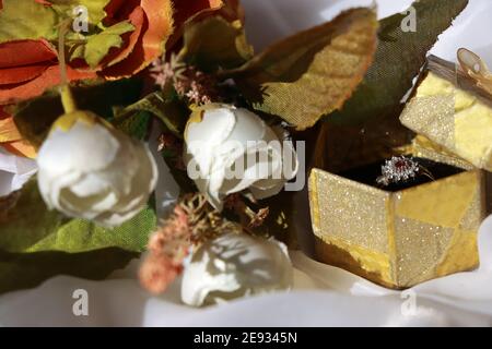 Verlobung und Ehe silberner Diamant Rubinring in einem goldenen Geschenkbox mit weißem Hintergrund und weißem und orangefarbenem Bouquet Stockfoto