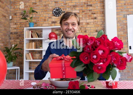 Lächelnder kaukasischer Mann, der einen Videoanruf macht und einen roten Haufen hält rosen und Geschenkbox Stockfoto