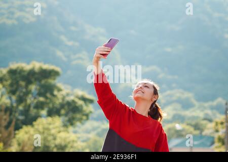 Videokommunikation, Blogging. Eine junge Frau posiert auf einer Telefonkamera, macht ein Selfie oder kommuniziert via Video-Link. Bäume und Berg auf dem Rücken Stockfoto