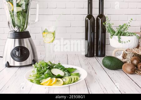 Mixer und Flaschen vor einer weißen Wand, Grün in einem Holzkoffer. Grüner Apfel, Zitrone, Rettich, Petersilie, Mangold, Minze, Marder, Avocado, Kiwi, Kohlr Stockfoto
