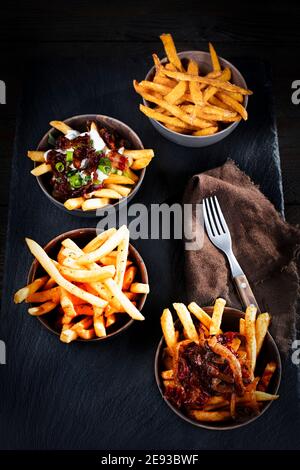 Vier verschiedene Pommes mit verschiedenen Sauce mit Zwiebel- oder Specksauce, in identischen Schüsseln auf einem schwarzen Skipperteller angeordnet Stockfoto