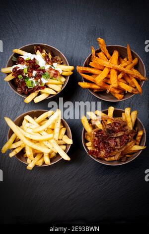 Vier verschiedene Pommes mit verschiedenen Sauce mit Zwiebel- oder Specksauce, in identischen Schüsseln auf einem schwarzen Skipperteller angeordnet Stockfoto