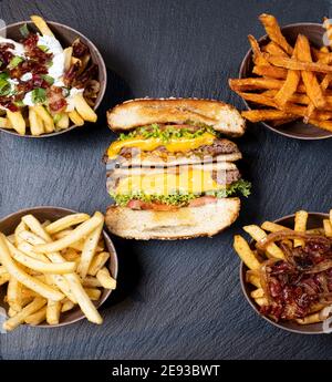 Vier verschiedene Pommes mit verschiedenen Sauce mit Zwiebel- oder Specksauce, in identischen Schüsseln auf einem schwarzen Skipperteller und einem Burger angeordnet Stockfoto