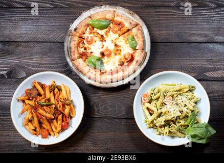 Rigatoni arrabiata, Rigatoni Pesto und eine Pizza auf einem braunen Holztisch mit Gabel und Messer Stockfoto