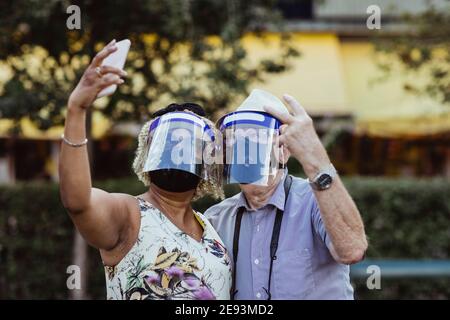 Ältere Frau, die Selfie auf einem Smartphone nimmt, das bei einem Mann steht In Park während COVID-19 Stockfoto