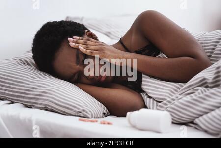 Kränkelnde junge afroamerikanische Dame mit geschlossenen Augen drückt die Hände Zu Tempeln und leidet unter Kopfschmerzen liegt auf dem Bett Stockfoto