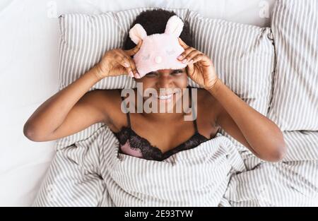 Glückliche junge afrikanisch-amerikanische attraktive weibliche Ausziehen rosa Schlaf Maske haben charmantes Lächeln Stockfoto