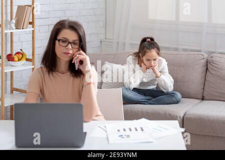 Beschäftigte Mutter, die online von zu Hause aus arbeitet, vernachlässigte Teenager-Tochter, die auf dem Sofa in der Nähe sitzt und unter Einsamkeit leidet Stockfoto