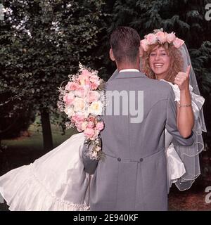 Hochzeit im Jahr 1989 von jungen Modell freigelassene Paar posiert im Freien Für Fotografien in 80s Mode Braut im Hochzeitskleid mit Blumenstrauß gibt Daumen nach oben getragen vom Bräutigam Rückansicht Grauer Morgenanzug und das Tragen einer lächelnden glücklichen Braut historischen 1980s Archiv Essex England Großbritannien Stockfoto