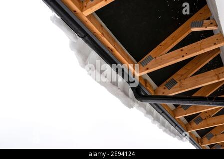 Das Dach eines Einfamilienhauses ist mit Schnee, sichtbaren Dachstühlen von unten und einer hängenden Schneekappe an der Rinne bedeckt. Stockfoto