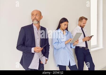 Gelangweilte, seriöse, mehrjährige Menschen, die sich im Wartebereich im Büro anstellen und Gadgets verwenden Stockfoto