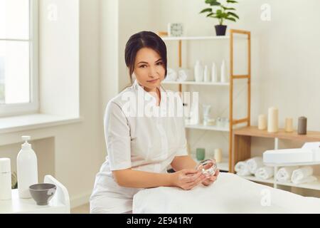 Lächelnde Frau Dermatologe sitzt, hält feuchtigkeitsspendende Maske im Glas und Blick auf die Kamera Stockfoto