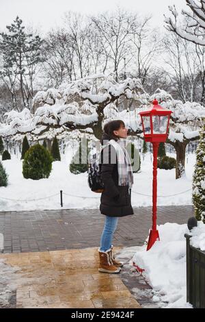 Junge lächelnde Frau, die in eine alte rote Laterne mit weihnachtsdekoration in einem schneebedeckten Winterpark schaut. Schneebedeckter Winter Stockfoto