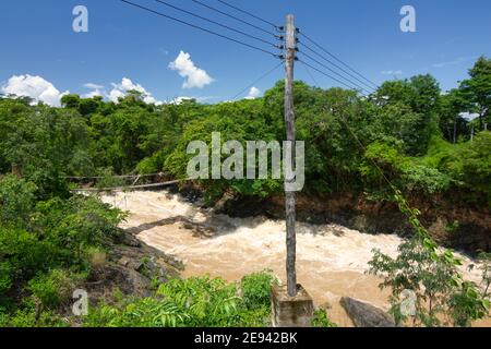 Laos, Bambusbrücke über schlammigen Fluss, hölzerne elektrische Pfosten und Dschungel. Stockfoto