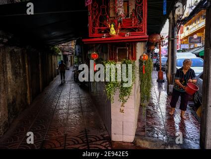 Eine ältere Thai-chinesische Frau gießt Wasser aus einem Eimer, um den Straßenbelag in der Chinatown-Gegend von Bangkok, Thailand, zu reinigen Stockfoto