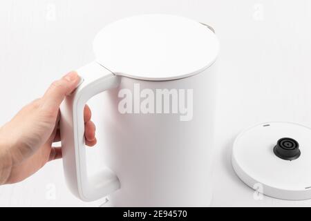 Eine weibliche Hand hält einen weißen Wasserkocher an der Griff auf einem weißen Holztisch Hintergrund Stockfoto