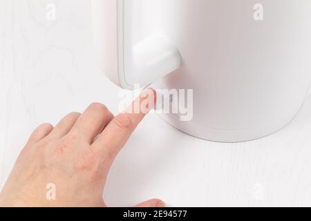 Eine weibliche Hand schaltet einen elektrischen weißen Wasserkocher an Wasser auf dem Hintergrund eines weißen Holztisches kochen Stockfoto