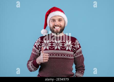 Positiver junger Mann in Urlaubspullover und Weihnachtsmütze, der Daumen hoch zeigt, lächelt und auf blauem Hintergrund auf die Kamera schaut Stockfoto