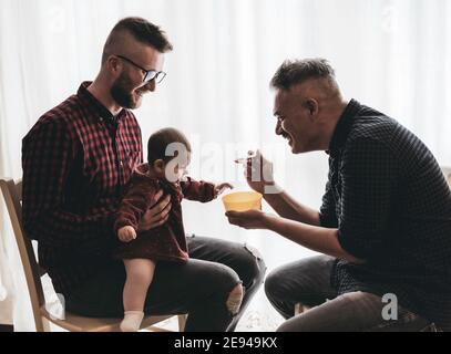 Männlich Gay paar mit angenommen Baby Mädchen zu Hause - Zwei schöne Väter füttern das Baby Mädchen in der Küche - Männliche Babysitter - LGBT Familie zu Hause - Vielfalt Stockfoto