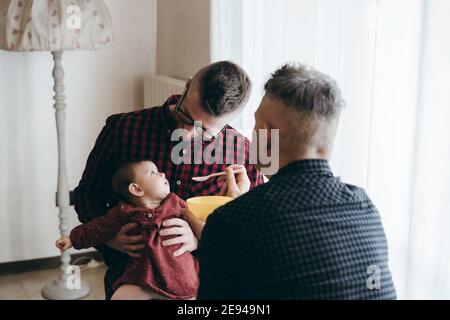 Männlich Gay paar mit angenommen Baby Mädchen zu Hause - Zwei schöne Väter füttern das Baby Mädchen in der Küche - Männliche Babysitter - LGBT Familie zu Hause - Vielfalt Stockfoto