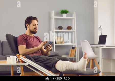 Junger Mann mit gebrochenem Bein sitzt in bequemen Sessel und Mit dem Mobiltelefon Stockfoto