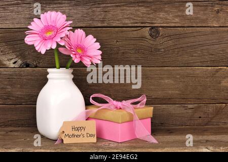 Happy Mothers Day Tag mit Geschenkbox und Vase von Rosa Blumen vor einem rustikalen Holzhintergrund Stockfoto