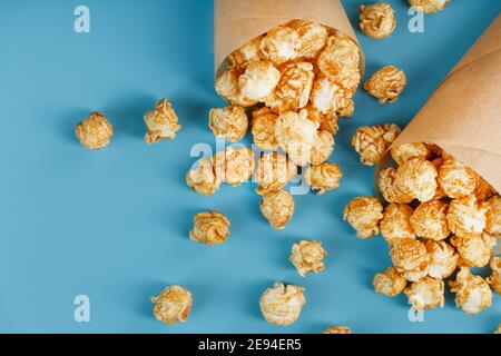 Karamell Popcorn in einem Papierumschlag auf blauem Hintergrund. Köstliches Lob für das Ansehen von Filmen, TV-Serien, Cartoons. Freier Speicherplatz, Nahaufnahme. Minimalisti Stockfoto