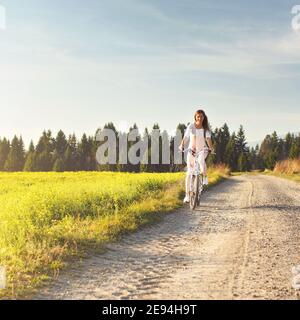 Junge Frau fährt Fahrrad auf staubiger Landstraße, Nachmittagssonne scheint auf sie, Blick von vorne Stockfoto