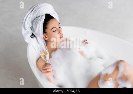 High-Angle-Ansicht der Frau bläst Schaum aus der Hand, während In der Badewanne mit Sektglas Stockfoto