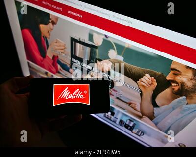 Person, die Mobiltelefon mit Logo des deutschen Kaffeehandelunternehmens Melitta auf dem Display vor der Website hält. Fokus auf Handy-Bildschirm. Stockfoto