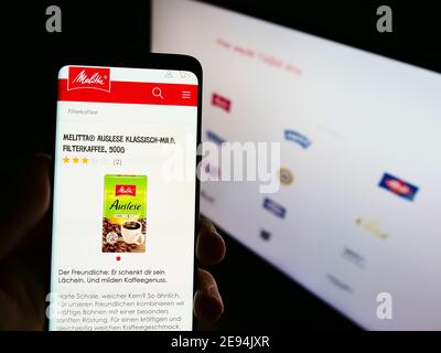 Person, die Smartphone mit Produkt und Logo des deutschen Kaffeehandelunternehmens Melitta auf dem Display mit Website hält. Konzentrieren Sie sich auf die Mitte des Telefonbildschirms. Stockfoto