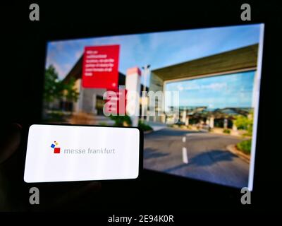 Person mit Smartphone mit Logo der Messe Frankfurt GmbH auf dem Display mit Webseite. Konzentrieren Sie sich auf die Mitte links auf dem Telefonbildschirm. Stockfoto