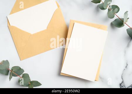 Basteln Sie Papierumschläge mit leeren Grußkarten Mockups und Eukalyptusblättern auf einem Marmortisch. Flach liegend, Draufsicht. Stockfoto