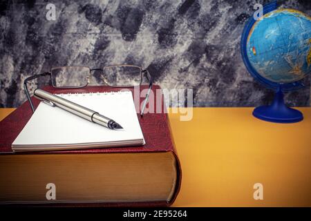 Auf dem Schreibtisch liegen ein großes Lexikonbuch und ein Notizbuch mit Stift. Artikel Buch und Brille, Globus und Notizbuch auf dem Tisch. Hintergrund mit Leerzeichen fo Stockfoto