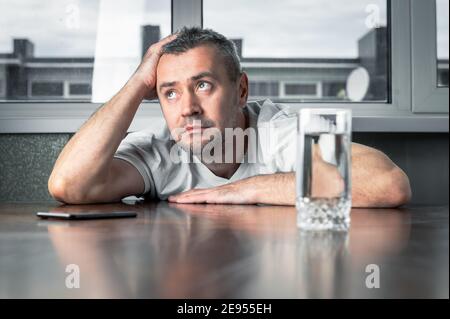 Gelangweilter junger Mann, der in seiner Küche sitzt und sein Smartphone hält. Stockfoto