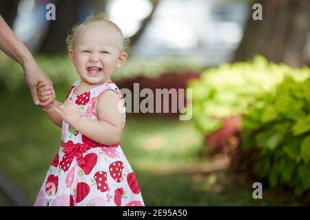 Ein kleines Mädchen von ein oder zwei Jahren im bunten Kleid hält die Hand eines Erwachsenen und lacht fröhlich. Drei Zähne im Mund. Nahaufnahme mit Platz Stockfoto