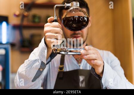 Der Barista schaut auf den Siphon mit Kaffee. Nahaufnahme alternativer Kaffeehähe. Kaffeebrühen mit Syphon Gerät im Café. Stockfoto