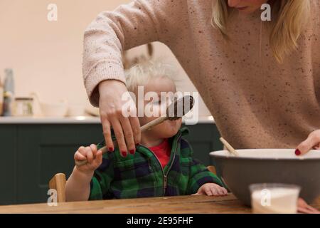 Netter kleiner Junge hilft, einen Kuchen mit Mama leckt einen Holzlöffel zu backen. Stockfoto