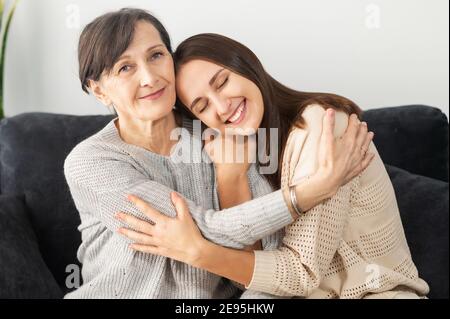Support und Liebe Konzept. Zwei Frauen unterschiedlicher Generationen umarmen sich, eine ältere Mutter und eine Erwachsene Tochter sitzen in Umarmungen auf der Couch und lächeln Stockfoto