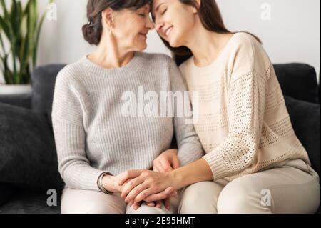 Eine ältere Mutter und eine Erwachsene Tochter sitzen von Angesicht zu Angesicht auf der bequemen Couch, eine Mutter und eine Tochter berühren die Stirn mit geschlossenen Augen. Familie Stockfoto