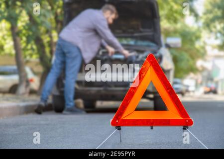 Mann, der Auto auf der Straße repariert. Fahrzeugpanne während der Fahrt. Der Fahrer sieht unter der Motorhaube des Fahrzeugs nach einer Panne. Rotes dreieckiges Stoppschild auf der Straße. Stockfoto