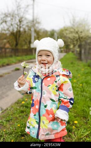 Ein kleines Mädchen in einer hellen Jacke lacht fröhlich draußen im Frühling und hält einen Zweig mit frühlingsweißen Blüten. Kinderglück. Stockfoto