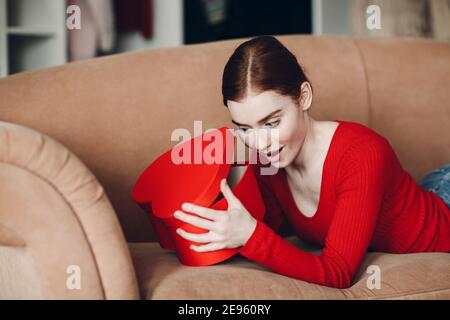 Schöne junge Frau mit roten Haaren liegt in ihrem Sofa im Wohnzimmer und öffnet die Geschenkbox in Form von Herz und Lächeln. Valentinstag Stockfoto