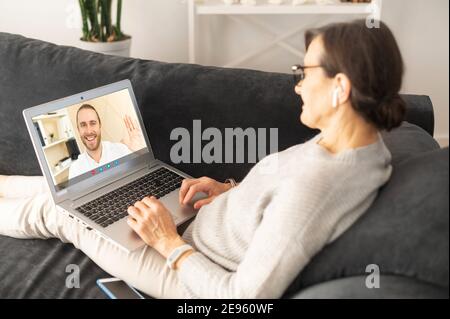 Eine lächelnde ältere Frau sitzt im Wohnzimmer, entspannt sich und spricht mit ihrem erwachsenen Sohn auf dem Laptop-Bildschirm per Videoanruf Stockfoto