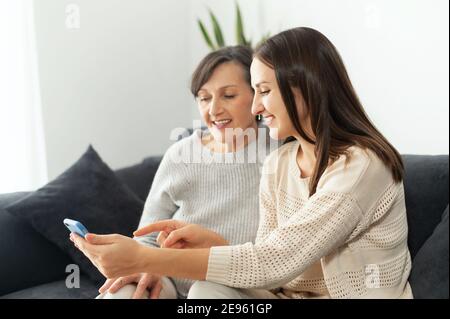 Eine Erwachsene Tochter hilft der Mutter herauszufinden, wie man ein Smartphone benutzt, zwei Generationenfrauen sitzen auf der Couch und schauen auf den Bildschirm des Telefons Stockfoto