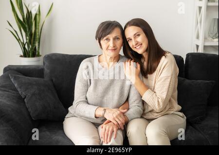 Ein Porträt einer älteren Mutter und einer erwachsenen Tochter, die auf der bequemen Couch sitzt, eine Mutter und eine Tochter schauen in die Kamera und lächeln umarmt. Familie Stockfoto