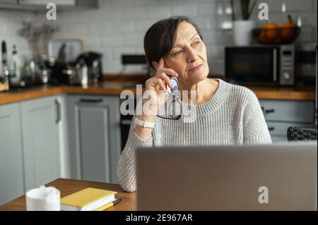 Nachdenkliche Rentnerin, die einen Laptop am Schreibtisch in der Küche für Fernarbeiten benutzt. Eine ältere Geschäftsfrau, Online-Lehrerin mit Computer Stockfoto
