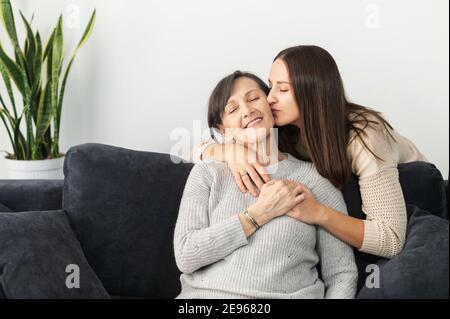 Eine Erwachsene Tochter umarmt und küsst eine ältere ältere Mutter auf dem Sofa im gemütlichen Wohnzimmer sitzen, zwei verschiedene Generationen Frauen umarmt drinnen Stockfoto