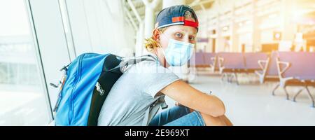 Porträt eines einsamen Teenager-Alleinreisenden mit Rucksack in leerer Flughafen-Passagiertransferhalle in schützender Gesichtsmaske und trauriger Betrachtung der Kamera. Tr Stockfoto