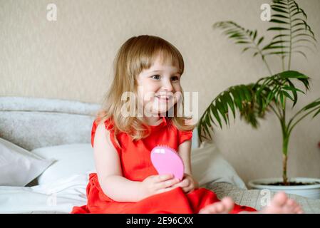 Niedliches rothaariges blauäugiges emotionales Mädchen lacht. Auf einem weißen Bett im Schlafzimmer. Mit einem Kamm in den Händen, einer rosa Haarbürste. 2 - 3 Jahre alt Stockfoto